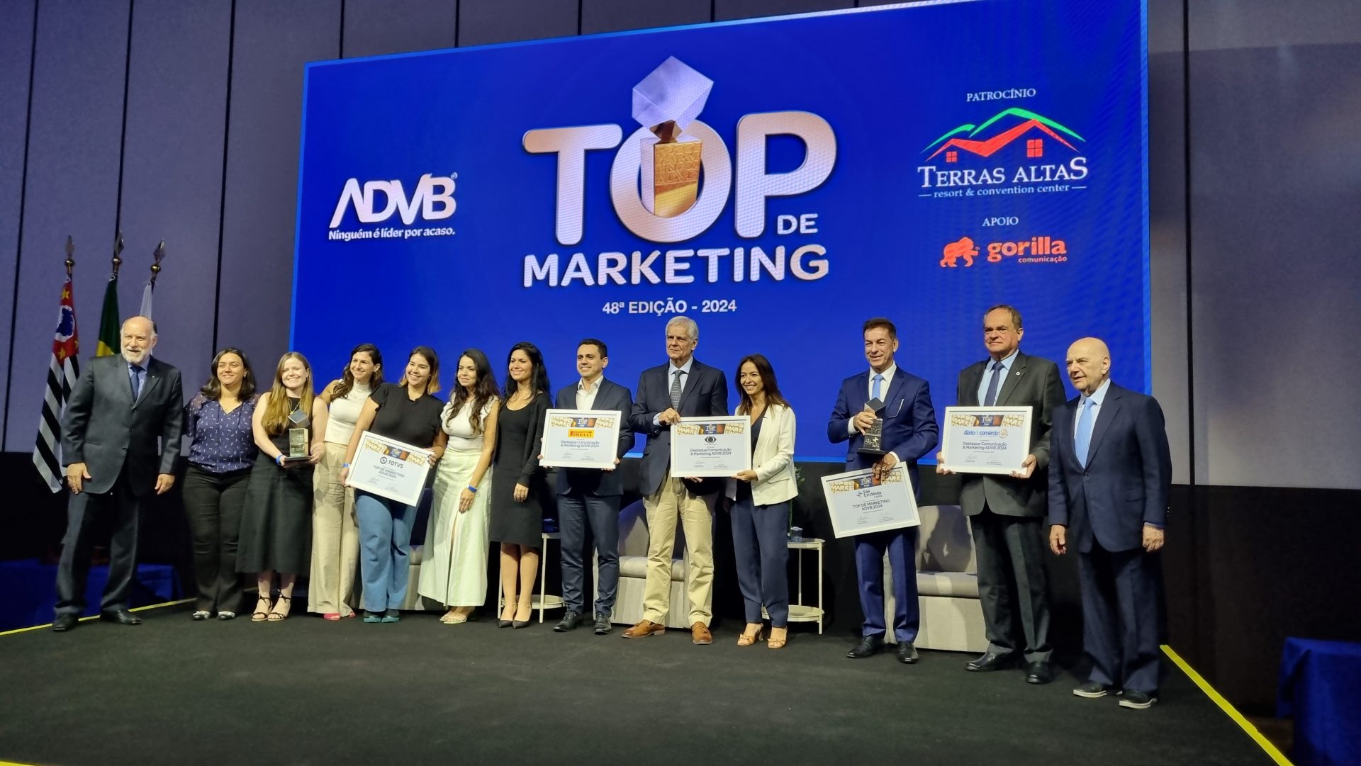 Grupo de premiados e organizadores posam em um palco durante a 48ª edição do prêmio Top de Marketing ADVB 2024. Ao fundo, uma grande tela azul com o logo da ADVB e a inscrição 'Top de Marketing 48ª Edição - 2024'. Algumas pessoas seguram troféus e certificados. À esquerda, bandeiras do Brasil e do estado de São Paulo.